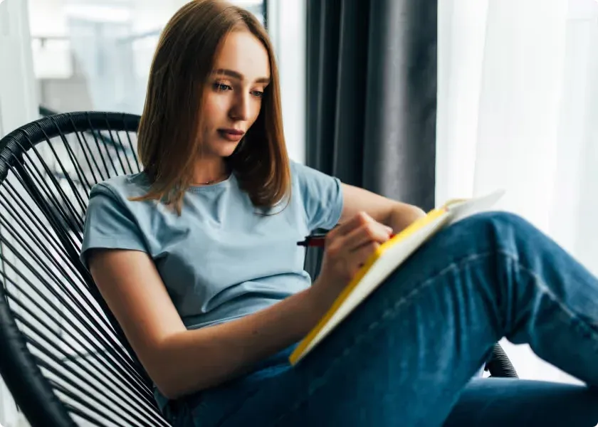 A young woman sitting in a rattan chair writes in a notebook.