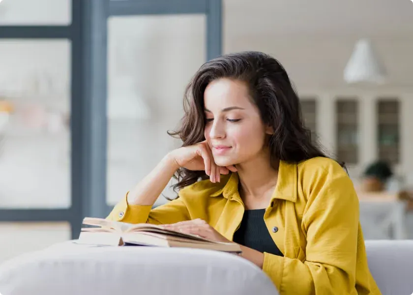A woman reads a book on a sofa beside a bright window.