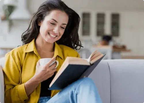 A smiling woman in a yellow shirt reads a hardcover on a sofa, a mug in her other hand.